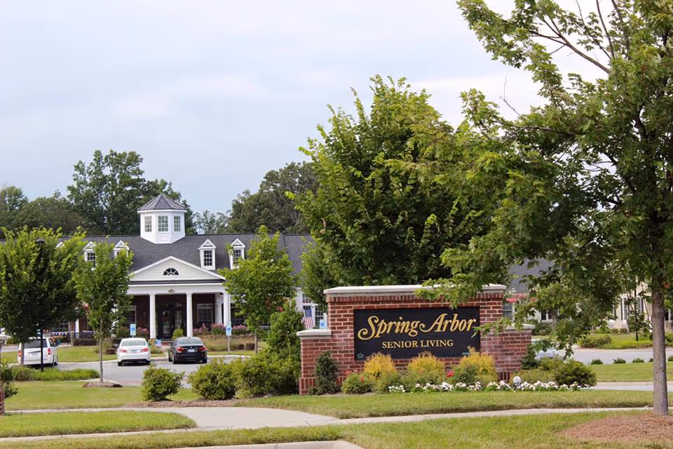 Brick entrance sign reading 'Spring Arbor Senior Living' in front of a large white senior living building surrounded by trees and parked cars.