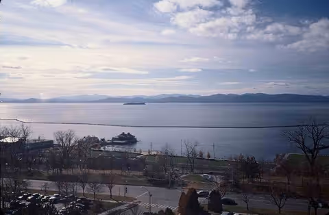 View of a large body of water with a ferry boat near the shore, trees and a road in the foreground, and distant mountains under a partly cloudy sky.