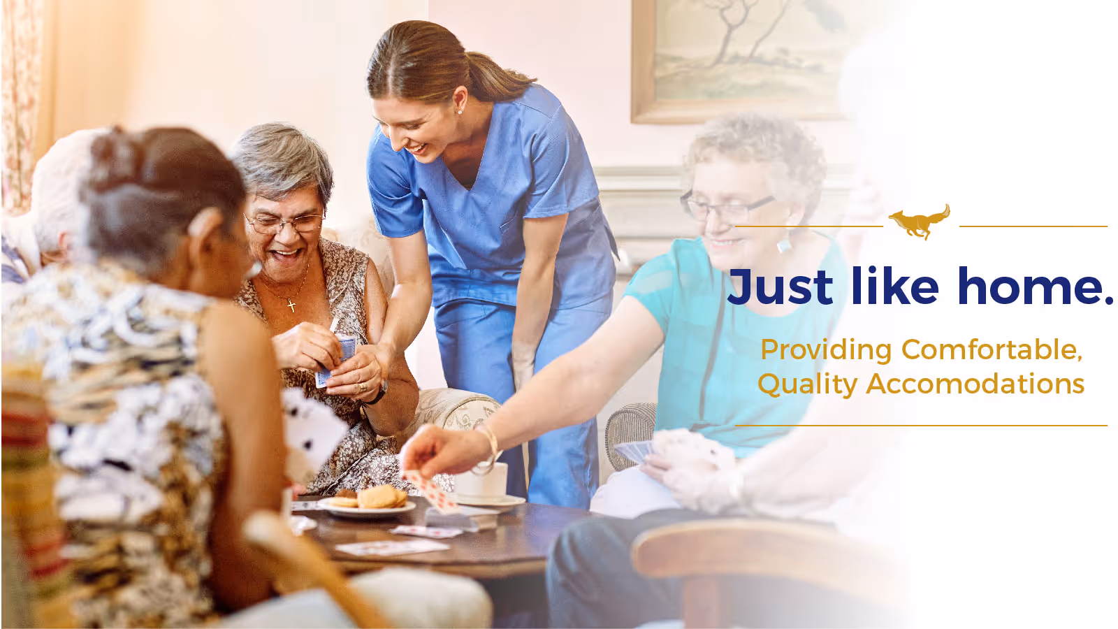 A group of elderly people sitting around a table playing cards and enjoying snacks, with a caregiver in blue scrubs smiling and engaging with them in a cozy living room setting.