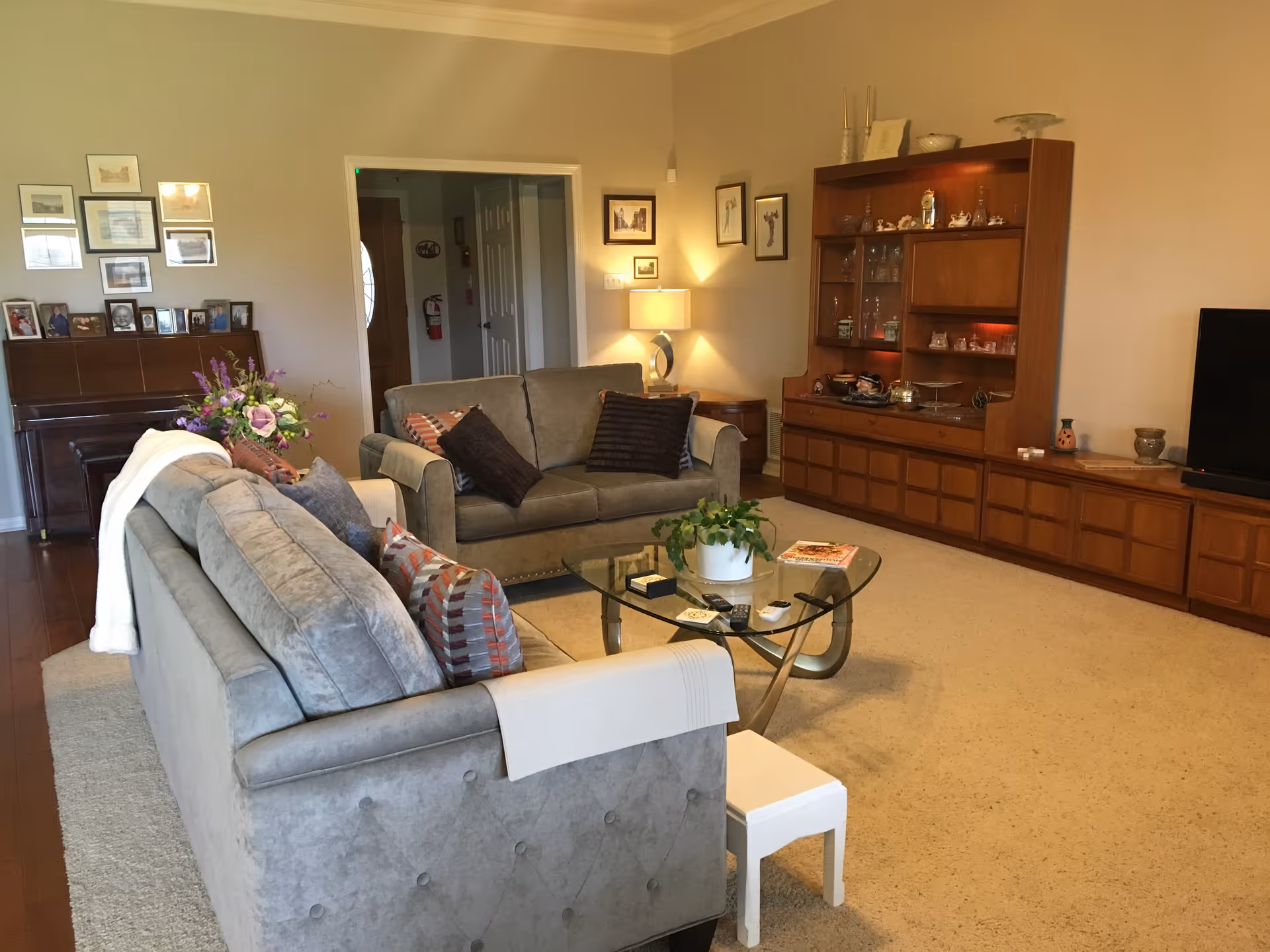 A cozy living room with two gray sofas adorned with various pillows and a white throw blanket. A glass coffee table with a plant and some magazines sits in the center on a beige carpet. Against the wall is a large wooden cabinet with shelves displaying decorative items and a flat-screen TV on the right. On the left side of the room, there is a piano with framed photos and artwork above it. The room is warmly lit with a table lamp and natural light from an unseen window.