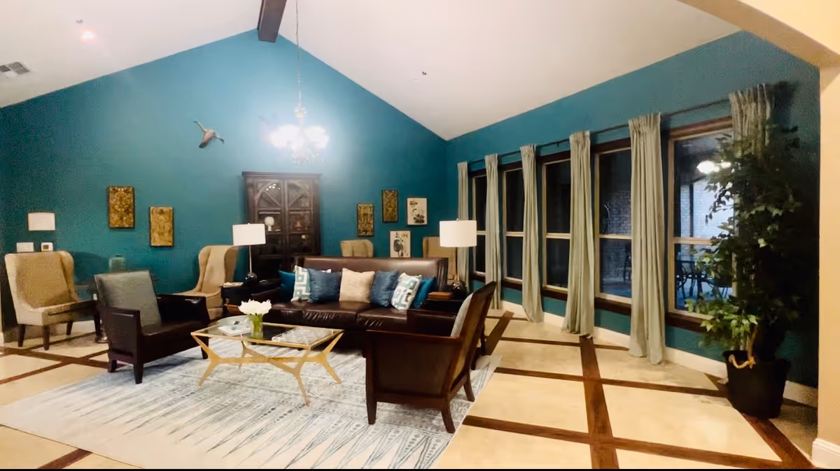 A spacious living room with teal walls and a high vaulted ceiling. The room features a dark brown leather sofa with multiple decorative pillows, two dark armchairs, and two beige wingback chairs. A glass coffee table with a gold frame sits on a patterned area rug. There are two table lamps on side tables flanking the sofa, a chandelier hanging from the ceiling, and several framed artworks on the wall. Large windows with light-colored curtains line one side of the room, and a tall potted plant is placed near the windows.