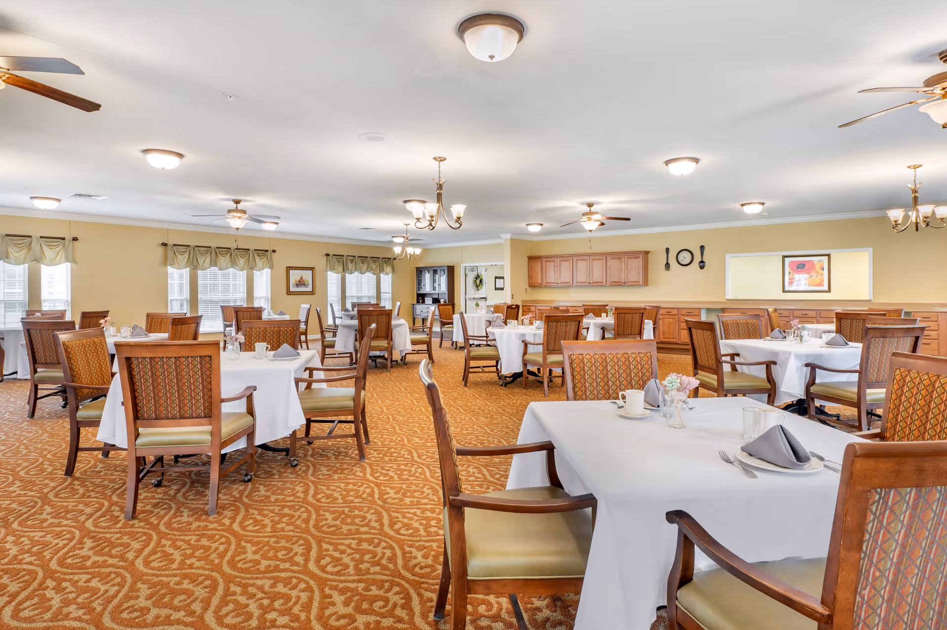 A spacious dining room with multiple tables covered in white tablecloths, each set with plates, napkins, and utensils. The room features patterned carpet, wooden chairs with cushioned seats, ceiling fans, and chandeliers. Large windows with valances allow natural light to fill the space, and there are cabinets and framed artwork on the walls.