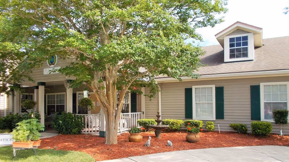 Exterior view of a single-story building with beige siding and green shutters, surrounded by landscaped bushes, potted plants, and a tree in front. There is a small fountain and two rabbit statues in the mulched garden area near the entrance, which has a covered porch with white railings.