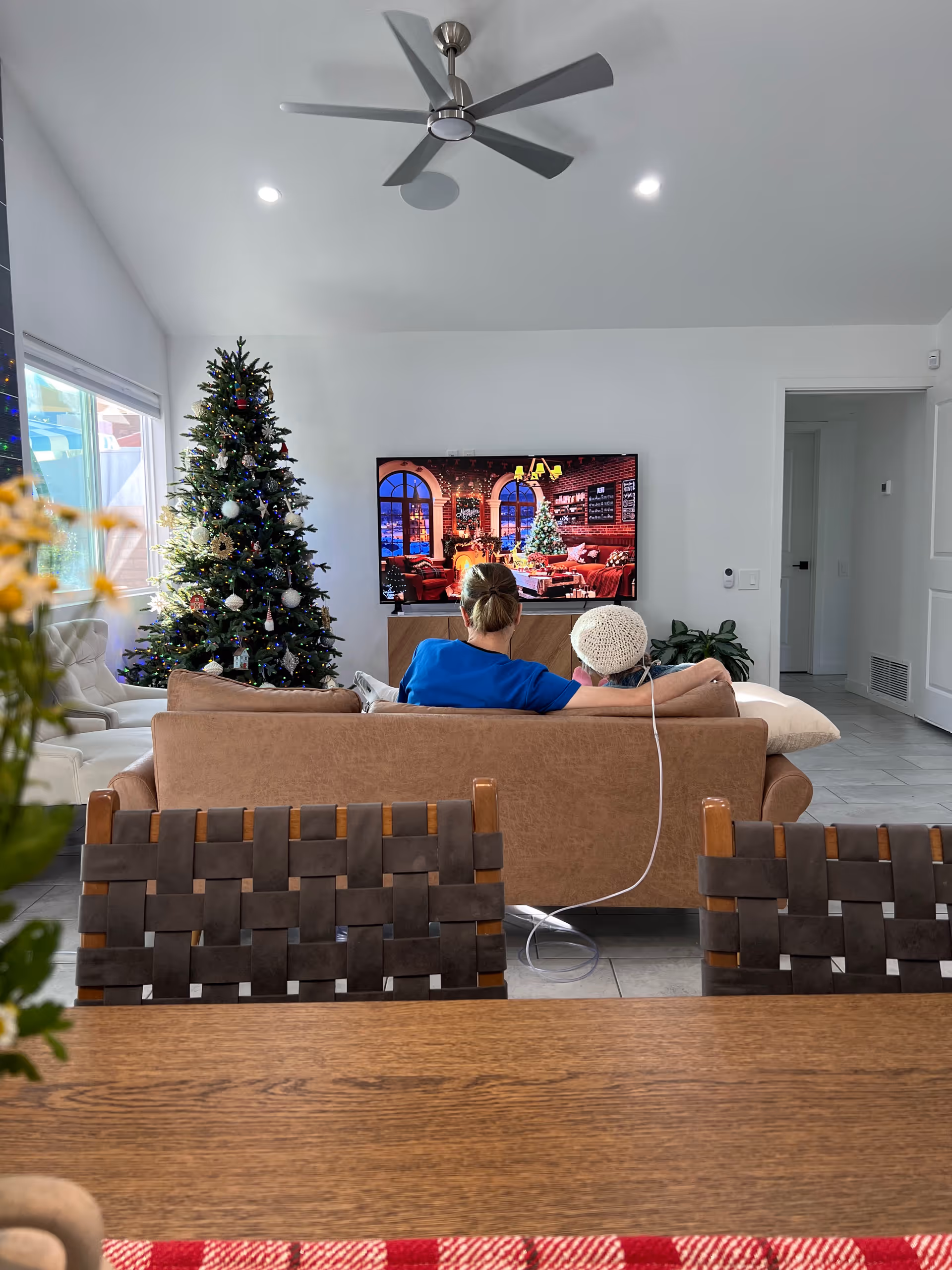 A cozy living room with a brown sofa facing a wall-mounted TV displaying a festive Christmas scene. A decorated Christmas tree stands to the left of the TV. Two people sit on the sofa, one wearing a blue shirt and the other wearing a white knit hat, with an oxygen tube connected to the person in the hat. The room has a ceiling fan, recessed lighting, and a large window letting in natural light.