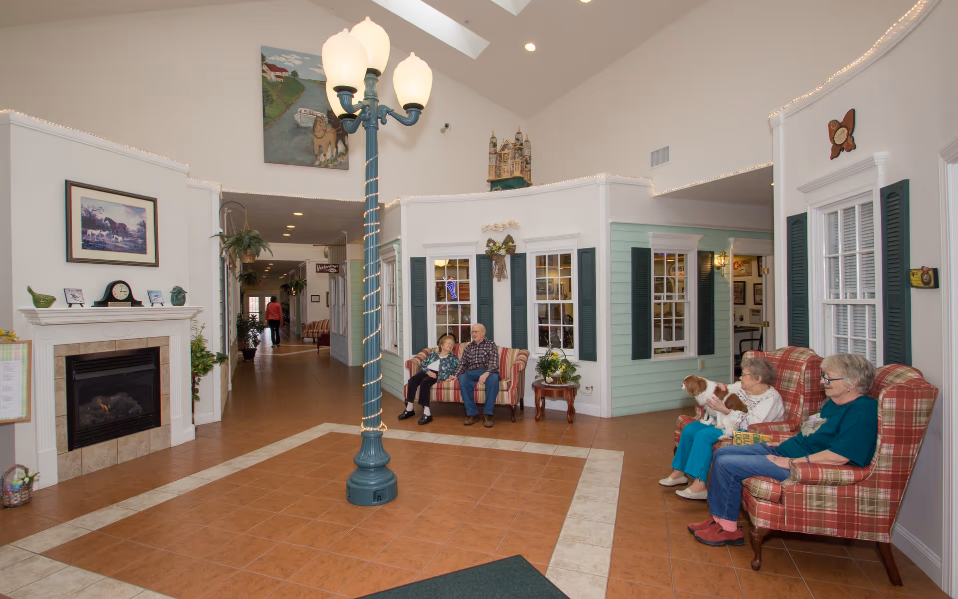A cozy common area in an assisted living community featuring a tall streetlamp-style light fixture in the center. There are two sets of plaid upholstered chairs and sofas where elderly residents are seated, one holding a small dog. The room has a fireplace with decorative items on the mantel, framed artwork on the walls, and a hallway leading further into the facility. The walls are painted in soft pastel colors with white trim and green shutters on the windows.