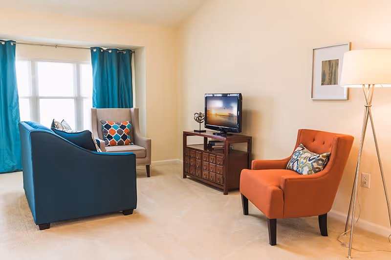 A cozy living room with a blue sofa, an orange armchair, and a beige armchair with colorful patterned pillows. There is a wooden TV stand with a flat-screen TV and a decorative item on it. The room has beige walls, a floor lamp, a framed picture on the wall, and large windows with teal curtains letting in natural light.