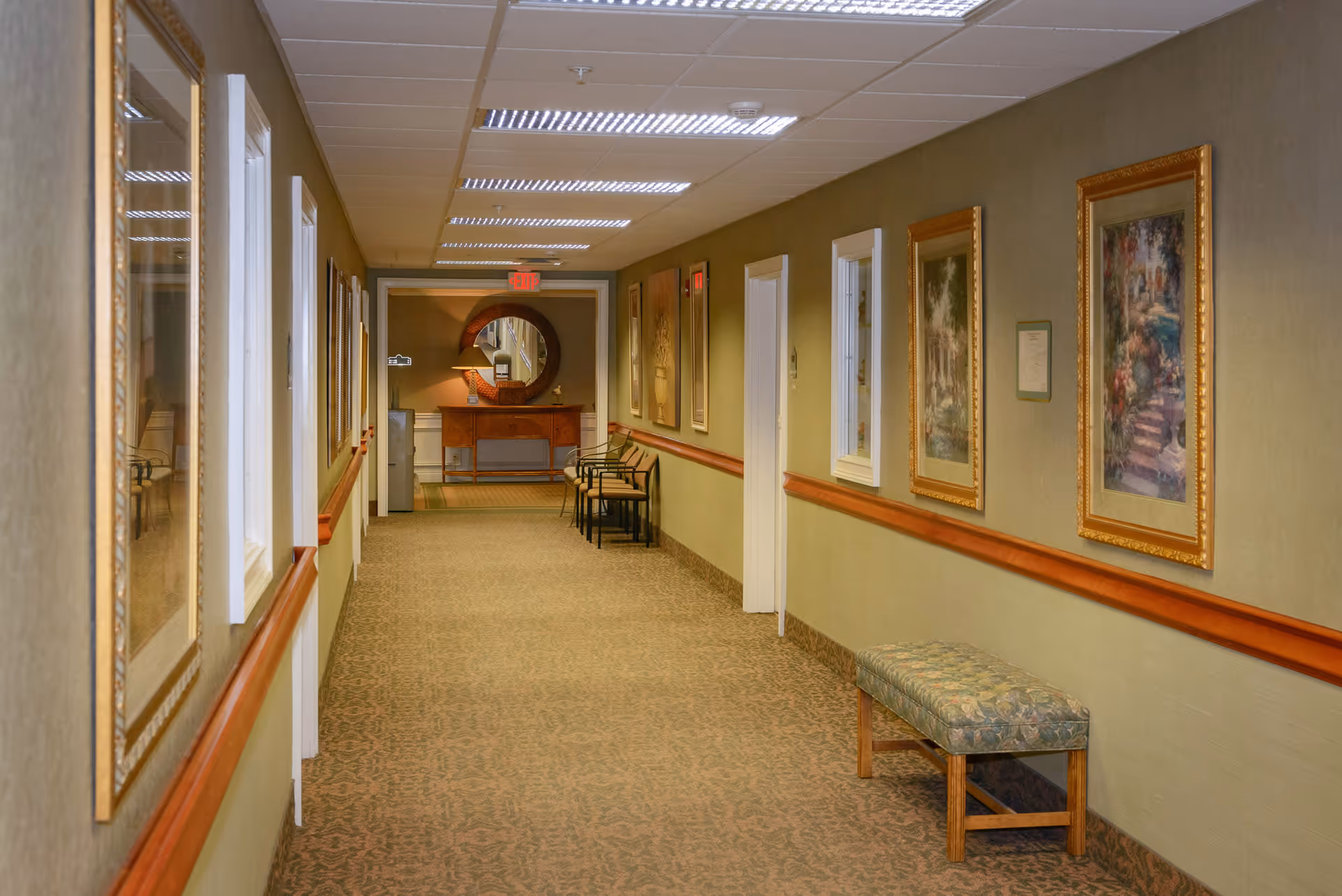 A well-lit hallway in a senior living facility with patterned carpet, framed artwork on the walls, a cushioned bench on the right side, and several chairs lined up near the end of the corridor. A wooden console table with a lamp and a round mirror is visible at the far end under an exit sign.