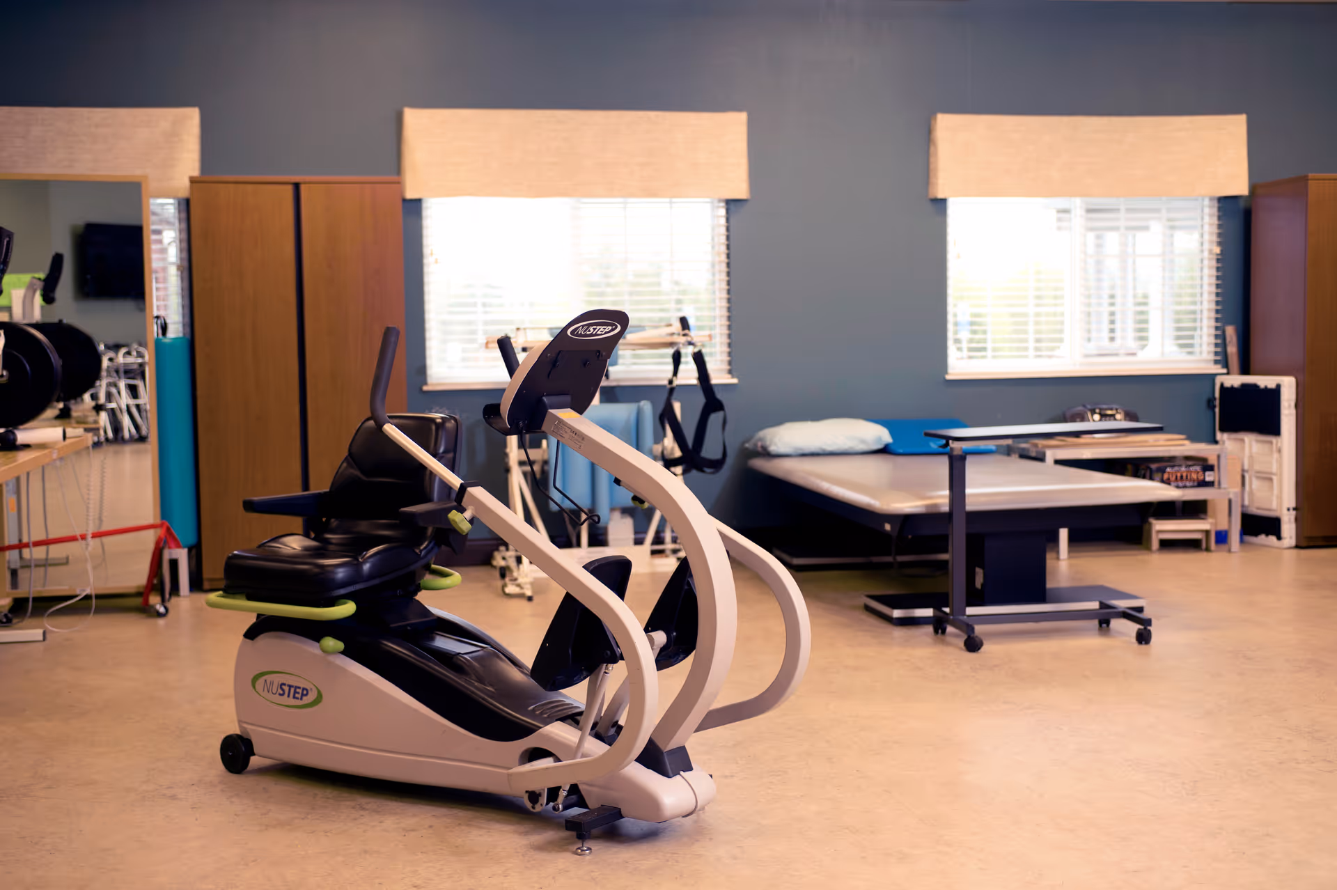 A rehabilitation room with exercise equipment including a NuStep recumbent cross trainer, therapy tables, and various physical therapy tools. The room has two windows with beige valances and blue walls.
