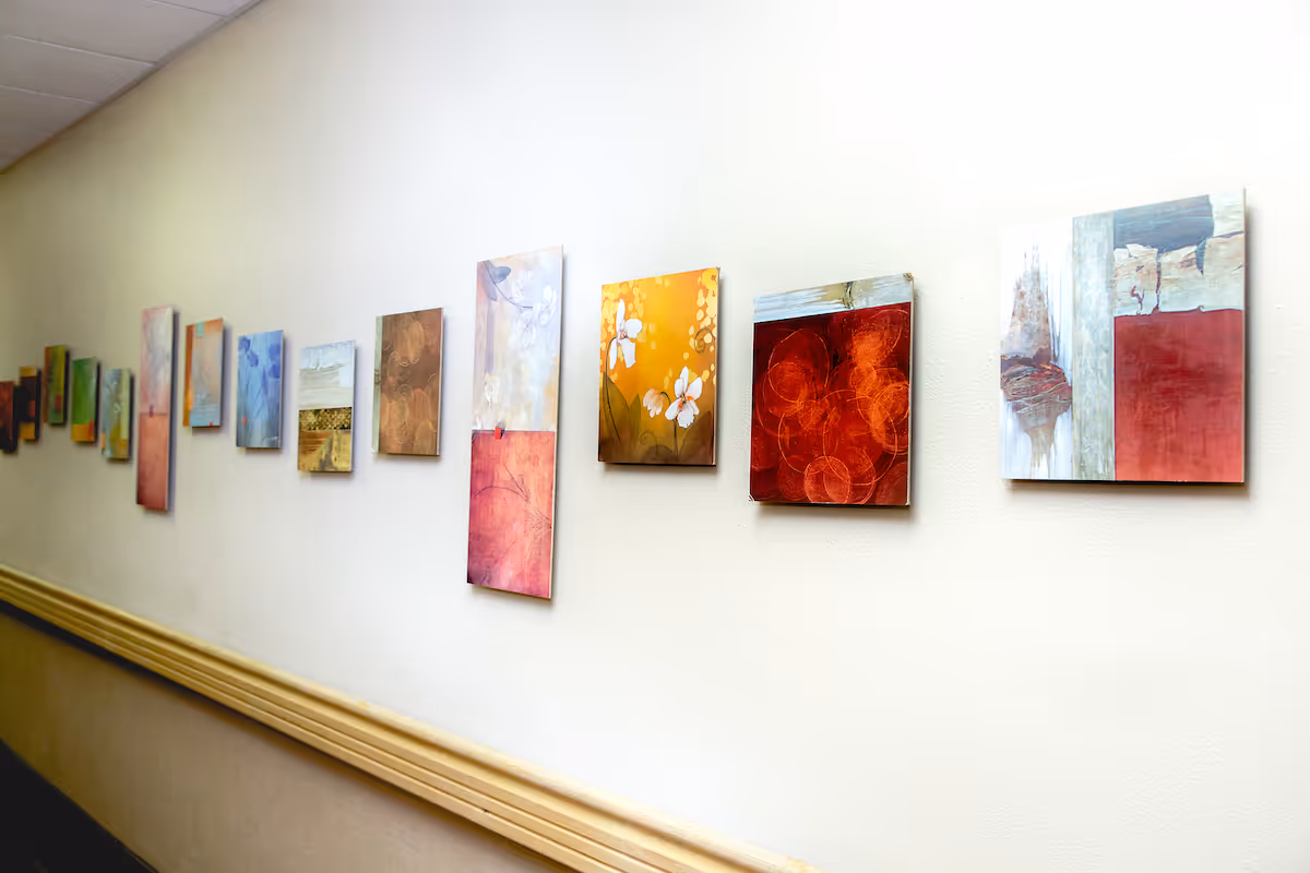 A hallway wall decorated with a series of small, colorful abstract paintings arranged in a horizontal line. The wall is light-colored and there is a wooden handrail along the lower part of the wall.