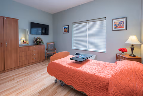 A tidy resident bedroom with a coral quilted bed, wooden dresser, wall-mounted TV, window with blinds, and a bedside table with a lamp.