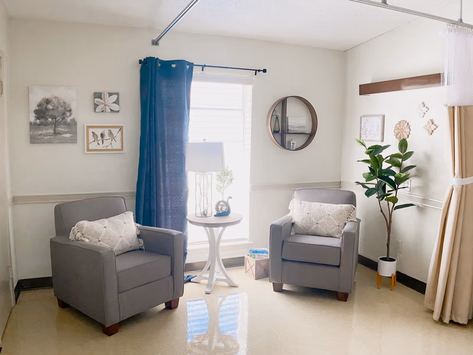 A cozy sitting area with two gray armchairs, each with a decorative pillow. Between the chairs is a small round white table with a lamp and a small plant. The room has a window with blue curtains and light walls decorated with framed artwork and a round wall shelf. There is also a tall potted plant in the corner and beige curtains on the right side.