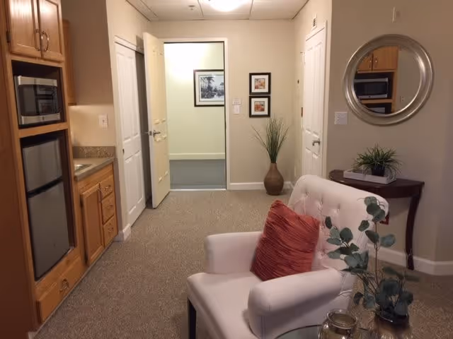 Interior view of a cozy assisted living facility room with a white armchair featuring a red pillow, a small wooden table with a plant and a round mirror above it, a kitchenette area with wooden cabinets, a microwave, and a mini refrigerator. The room has beige walls and carpeted floor, with an open door leading to another room decorated with framed pictures and a tall vase with greenery.