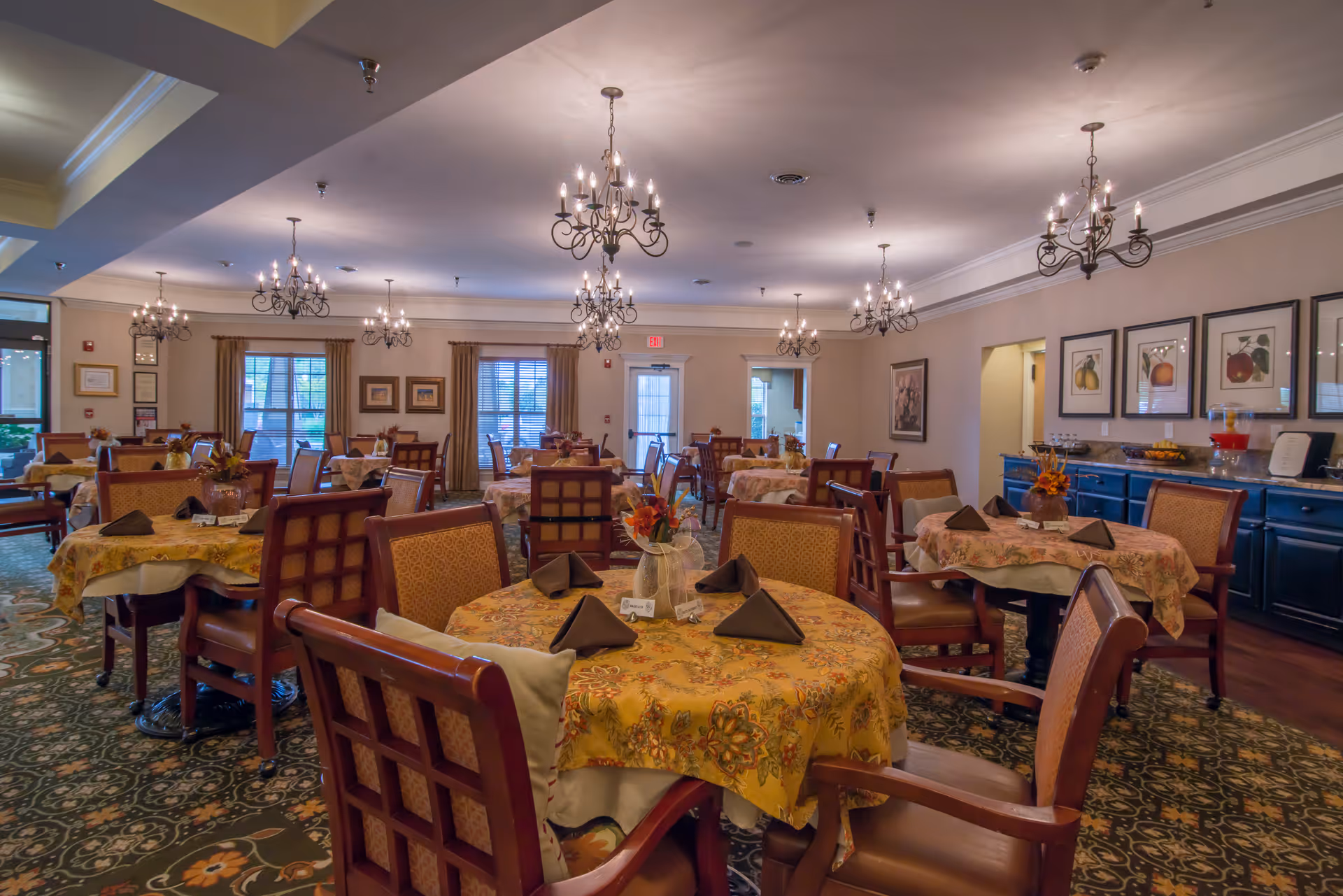 A spacious dining room with multiple round tables covered with floral tablecloths and set with folded napkins. The room features wooden chairs with cushioned seats, chandeliers hanging from the ceiling, framed artwork on the walls, and windows with curtains allowing natural light to enter.