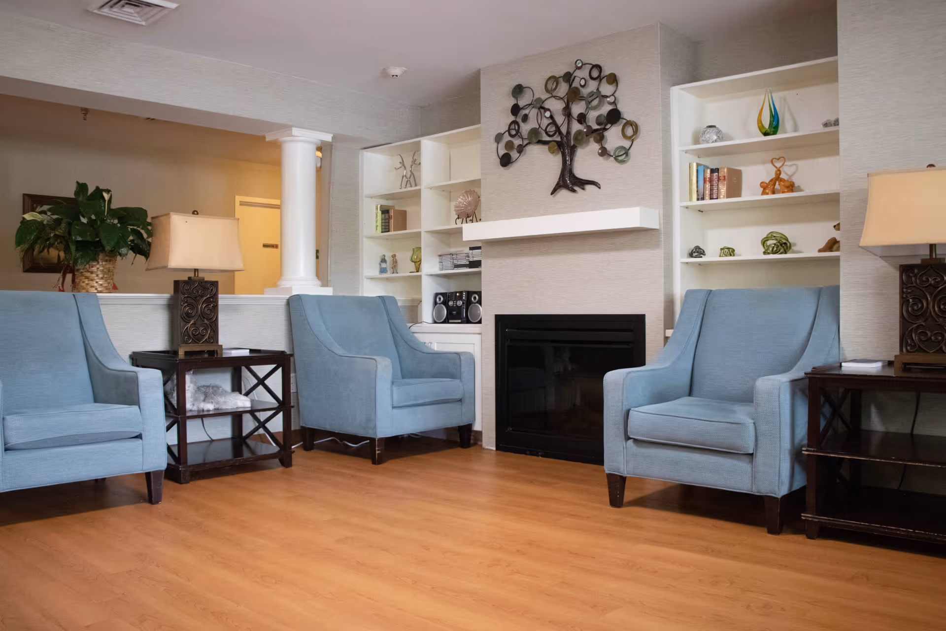 A cozy living room area with three light blue armchairs arranged around a black fireplace. The walls have built-in white shelves decorated with books, ornaments, and a stereo. There are two dark wooden side tables with lamps on either side of the chairs. A decorative metal tree wall art hangs above the fireplace. The floor is light wood, and a white column is visible in the background near a plant and a hallway.
