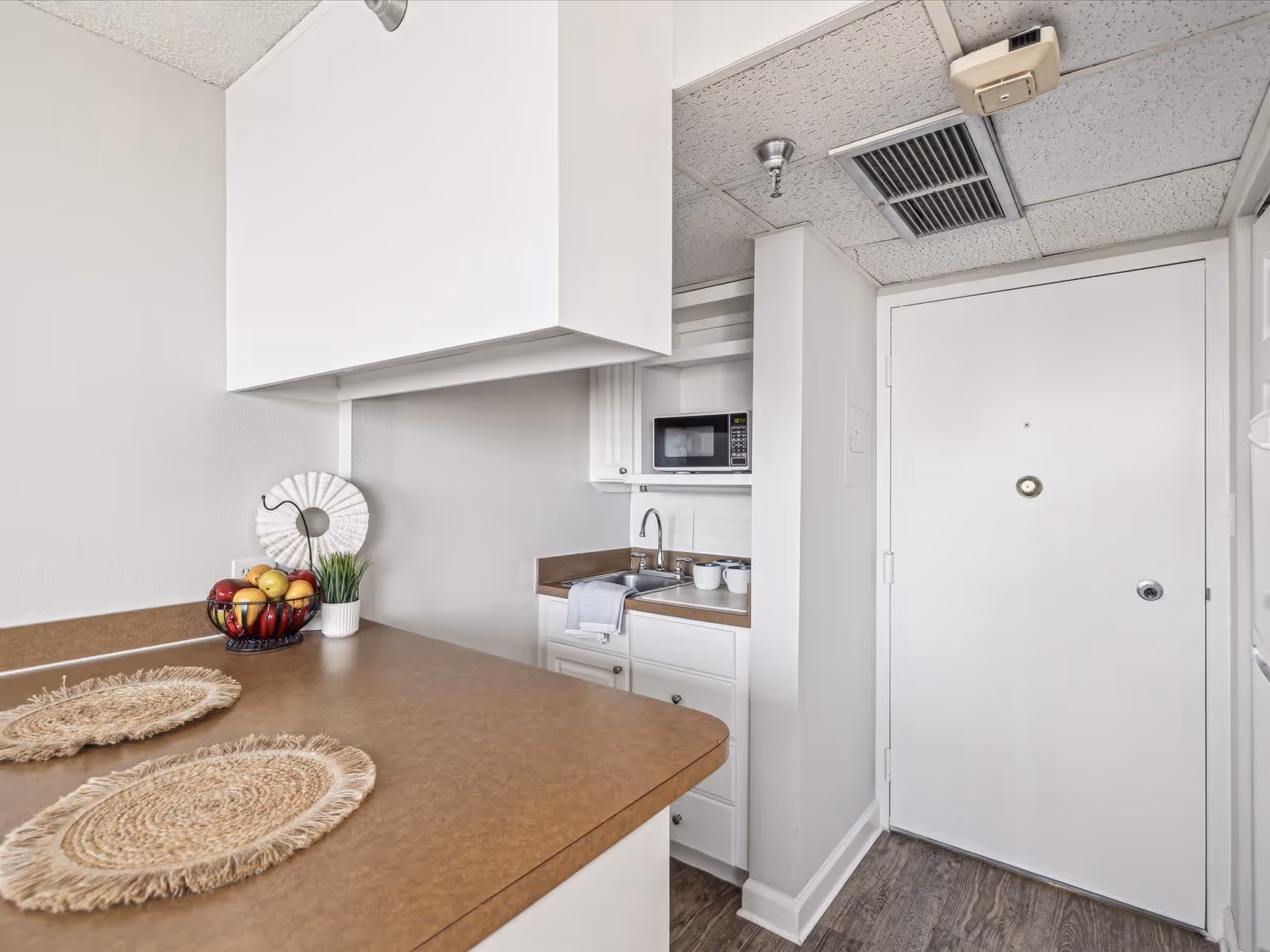 Small kitchen area with brown countertops, two woven placemats, a bowl of fruit, a decorative white circular object, and a small potted plant. White cabinets and a microwave above a sink with a towel hanging on the cabinet handle. A white door is visible on the right side with a peephole and lock.