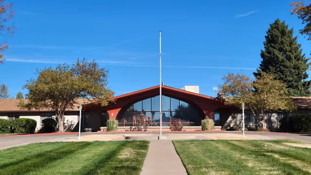 Front exterior view of Pueblo Heights Nursing and Rehabilitation building with a large triangular window, a flagpole in the center, surrounded by green grass, trees, and a clear blue sky.