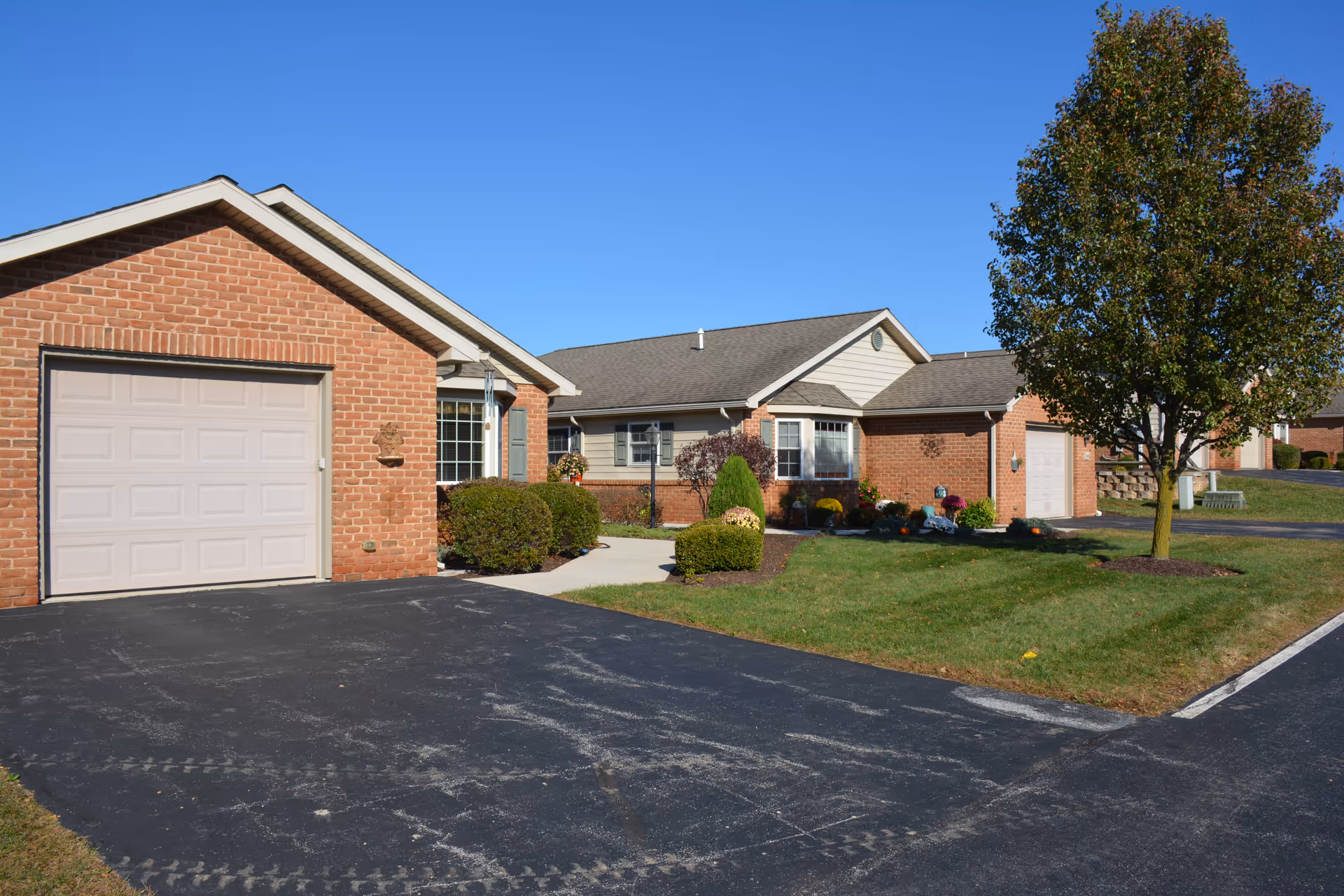 Exterior view of a single-story brick residential building with a garage, driveway, well-maintained lawn, bushes, and a tree under a clear blue sky.