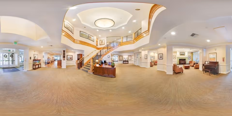 A spacious and well-lit senior living facility lobby with a central staircase leading to an upper floor. The area features comfortable seating, framed artwork on the walls, a piano, and a reception desk. Large windows and glass doors allow natural light to fill the space.
