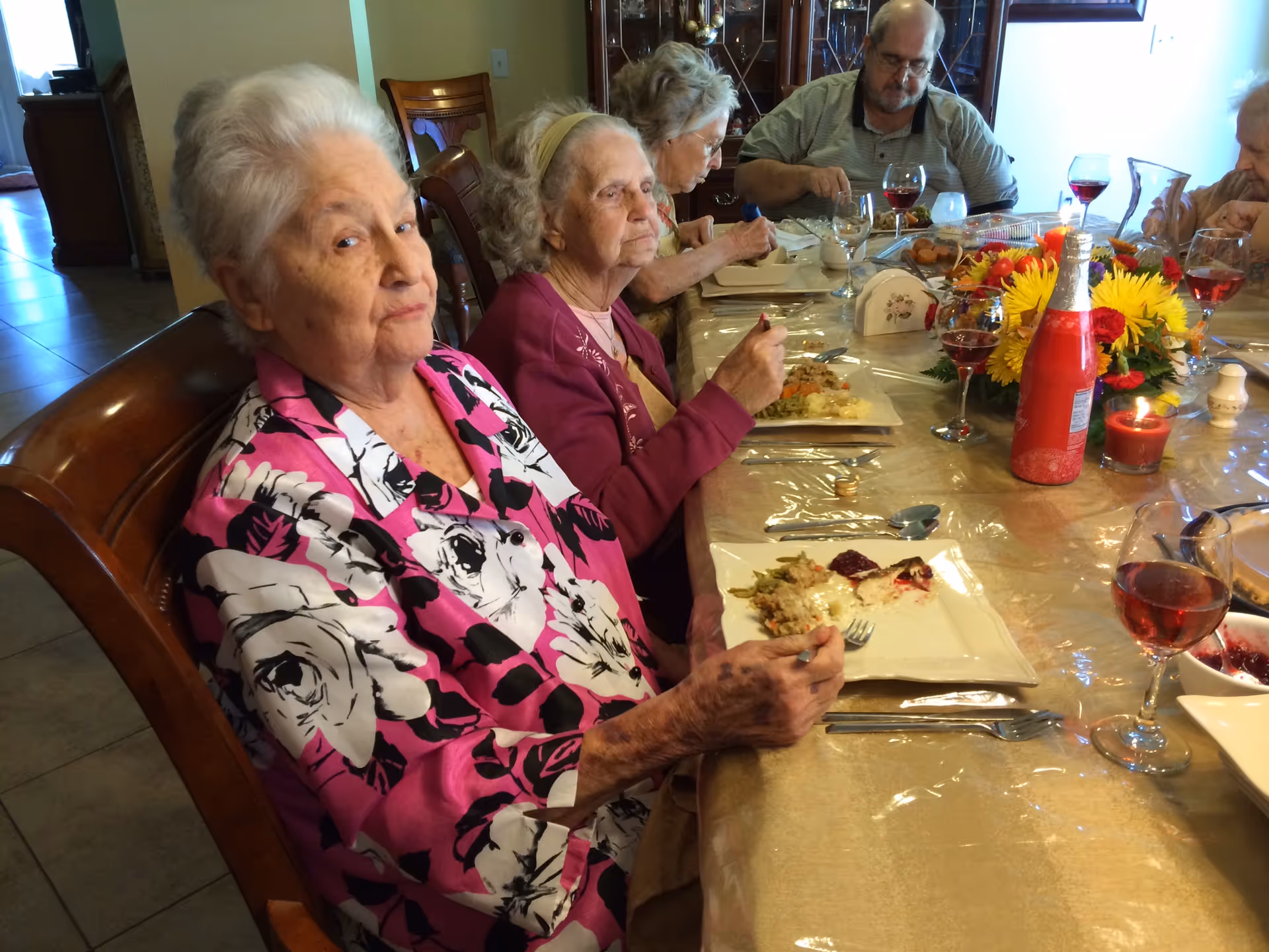 A group of elderly people sitting around a dining table enjoying a meal together. The table is set with plates of food, glasses of red wine, a floral centerpiece, and a lit candle. The setting appears warm and communal.
