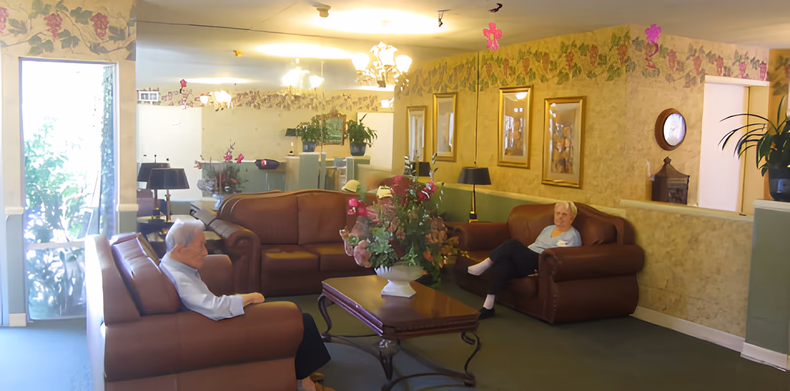 Two elderly residents seated on brown leather sofas in a decorated common living room with a central coffee table, floral arrangement, and wall art.