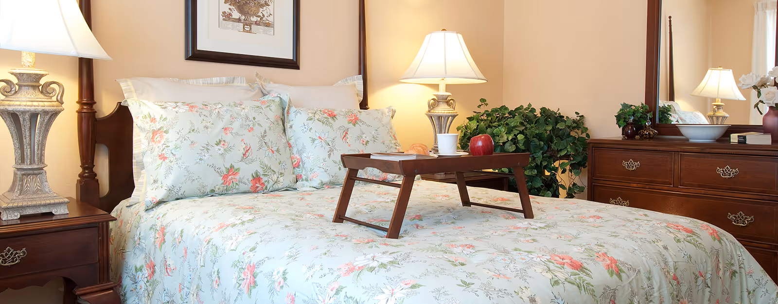 A cozy bedroom with a floral-patterned bedspread and matching pillows on a wooden bed frame. There is a wooden tray on the bed holding a cup, an apple, and a book. Two ornate table lamps with white shades sit on wooden nightstands on either side of the bed. A wooden dresser with a mirror and decorative items, including a plant and a vase with flowers, is visible in the background.
