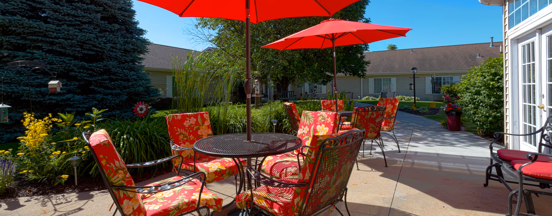 Outdoor patio with red floral chairs, tables, and large red umbrellas in a senior living courtyard.