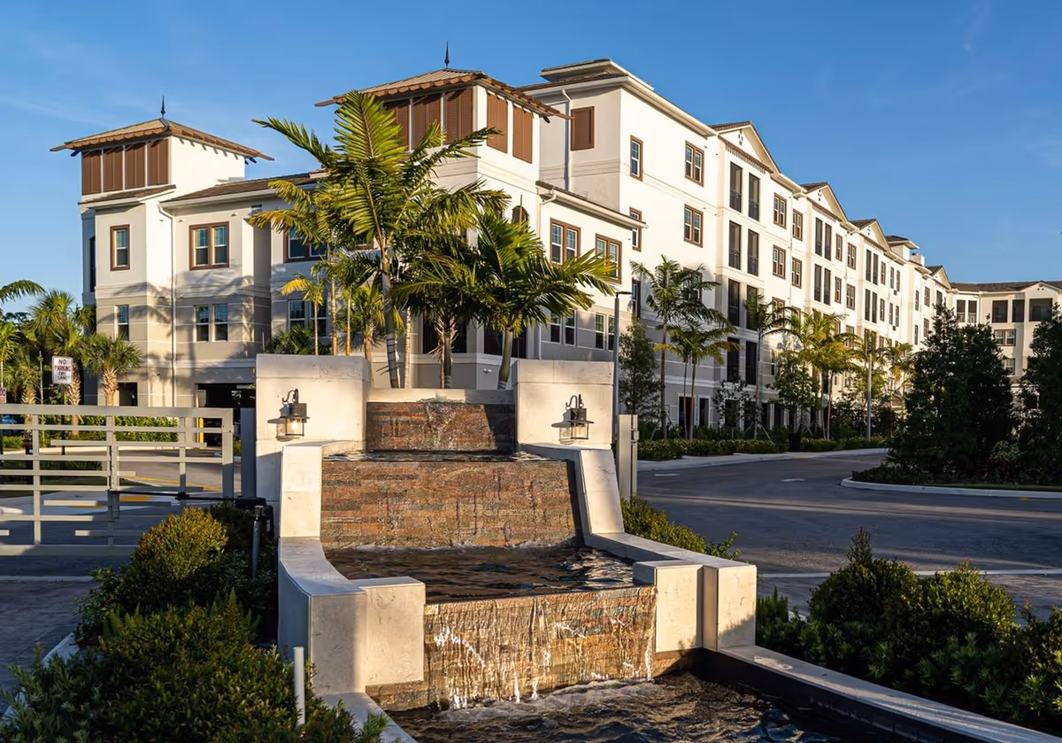 Exterior view of a multi-story residential building with white walls and brown window shutters, surrounded by palm trees and greenery. In the foreground, there is a tiered water fountain with water cascading down stone steps. The sky is clear and blue.