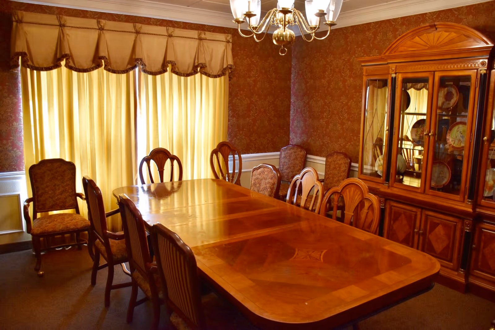A formal dining room with a large polished wooden dining table surrounded by eight upholstered chairs. The room features patterned wallpaper, a wooden china cabinet displaying plates, a chandelier hanging from the ceiling, and yellow curtains covering the windows.