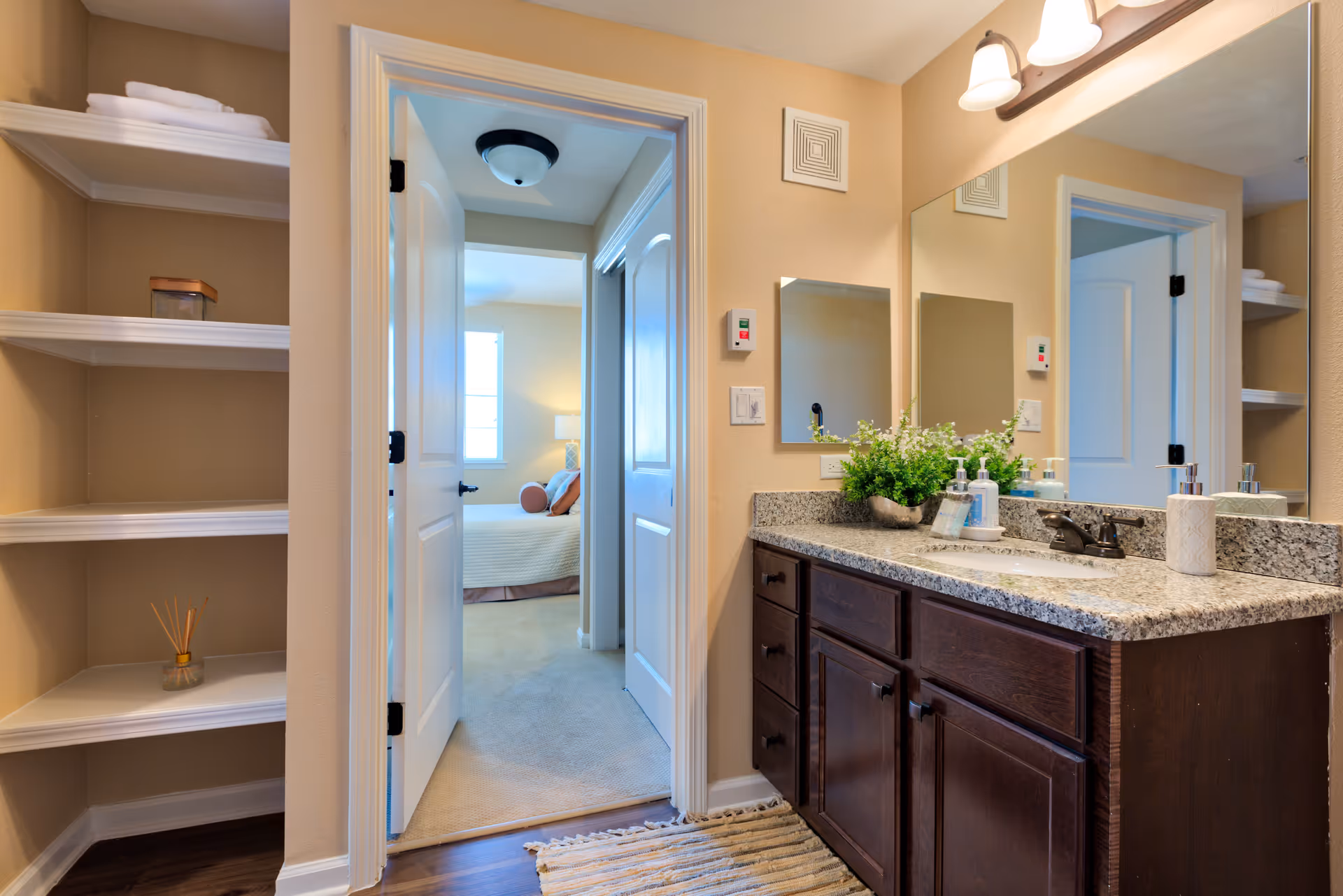 A bathroom with a granite countertop, dark wood cabinets, a sink, and a large mirror above. On the countertop are soap dispensers and a small plant. To the left, there are built-in white shelves with towels and a small decorative item. Through an open door, a bedroom with a bed and pillows is visible.