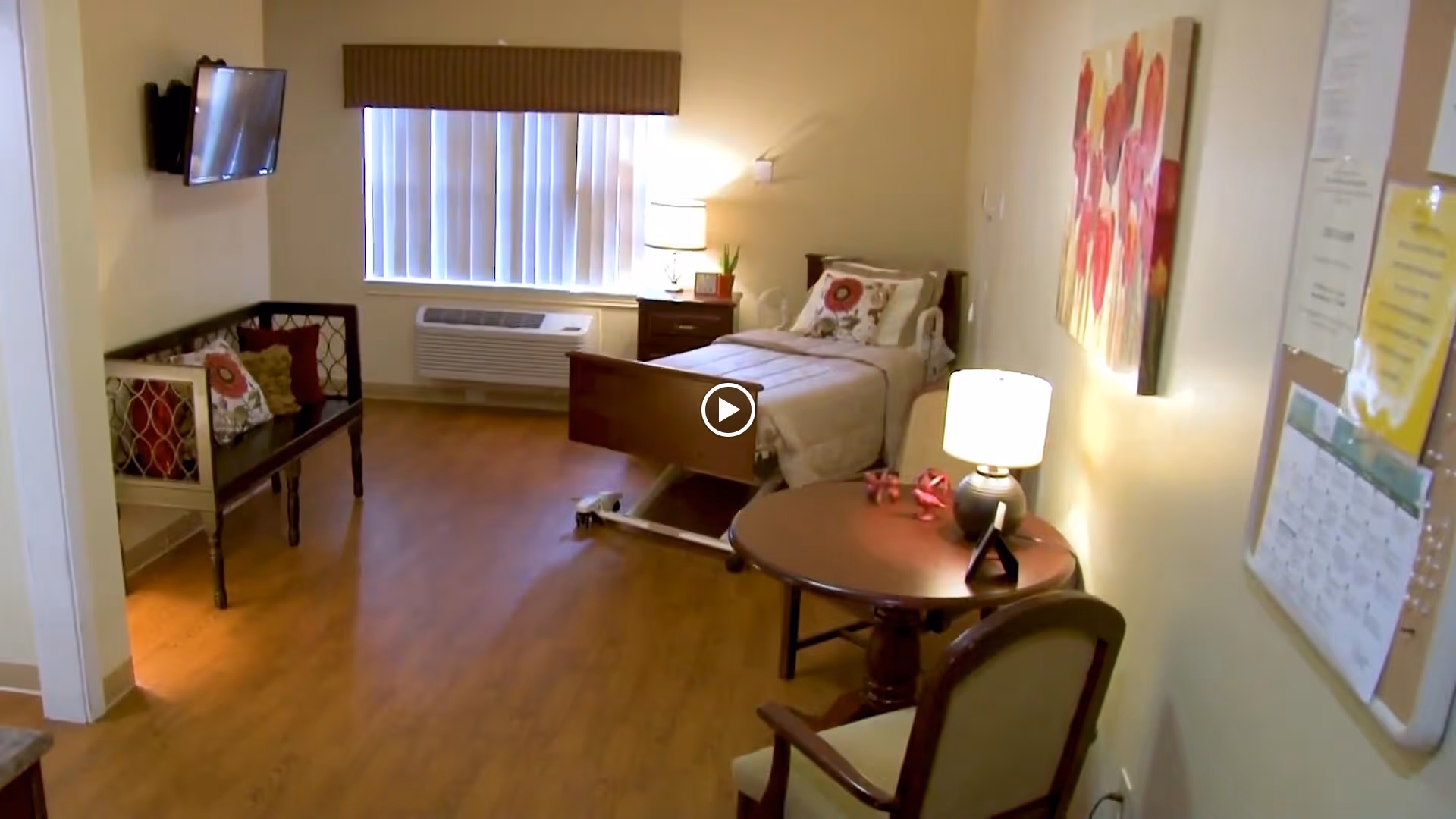 A cozy senior living room with a single bed against the far wall, a nightstand with a lamp and plant, a wall-mounted TV, a bench with decorative pillows, a round table with a lamp and chair, and a bulletin board on the wall. The room has wooden flooring and a window with vertical blinds.