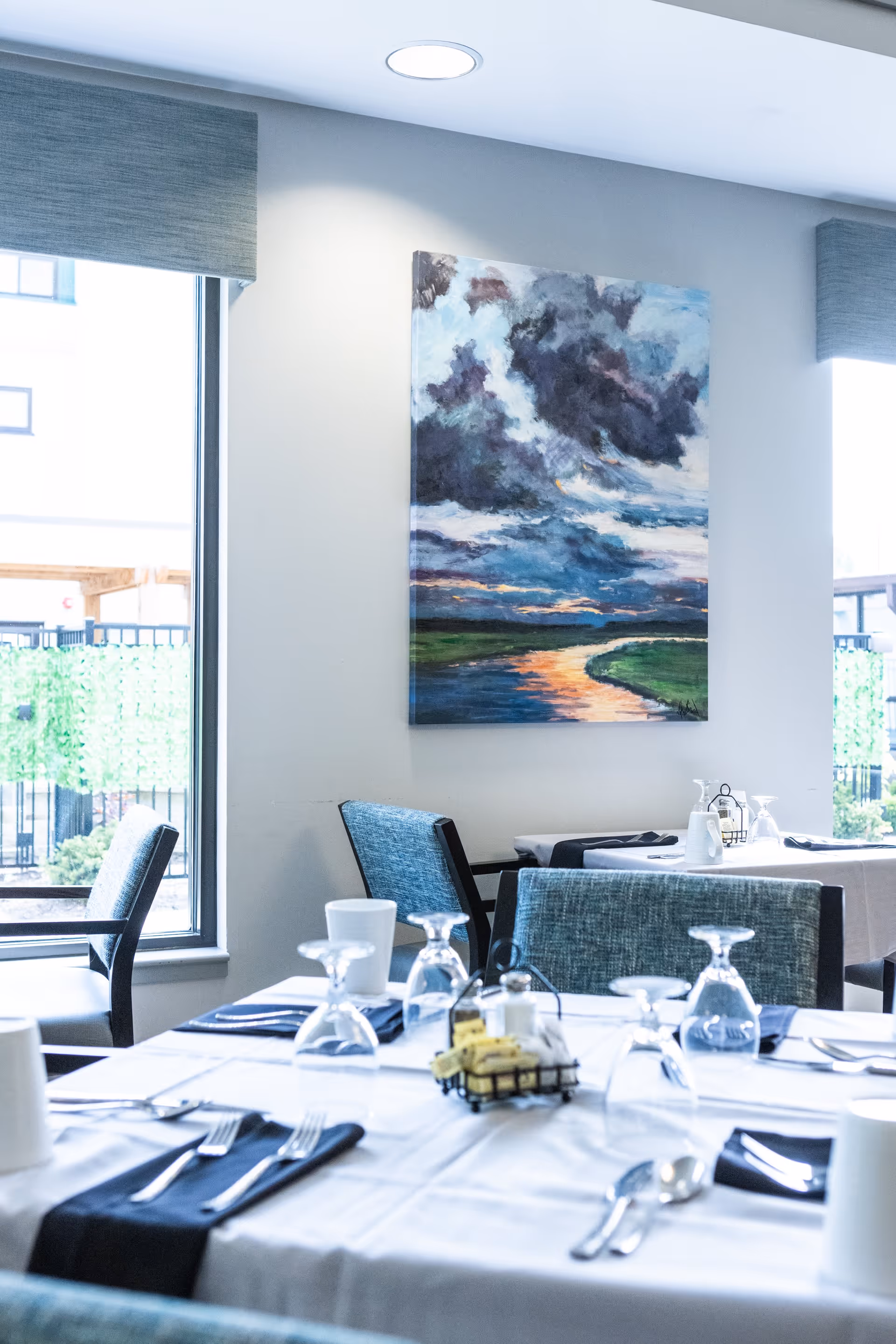 A dining room with tables set with white tablecloths, silverware, glasses, and napkins. There are blue upholstered chairs around the tables. A large window lets in natural light, and a colorful painting of a cloudy sky over a river hangs on the wall.