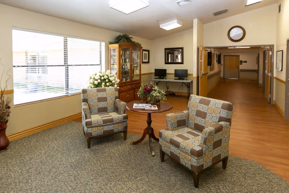 Sunlit seating area in a senior living facility with two patterned armchairs around a small table, a display cabinet, computer stations and a hallway.