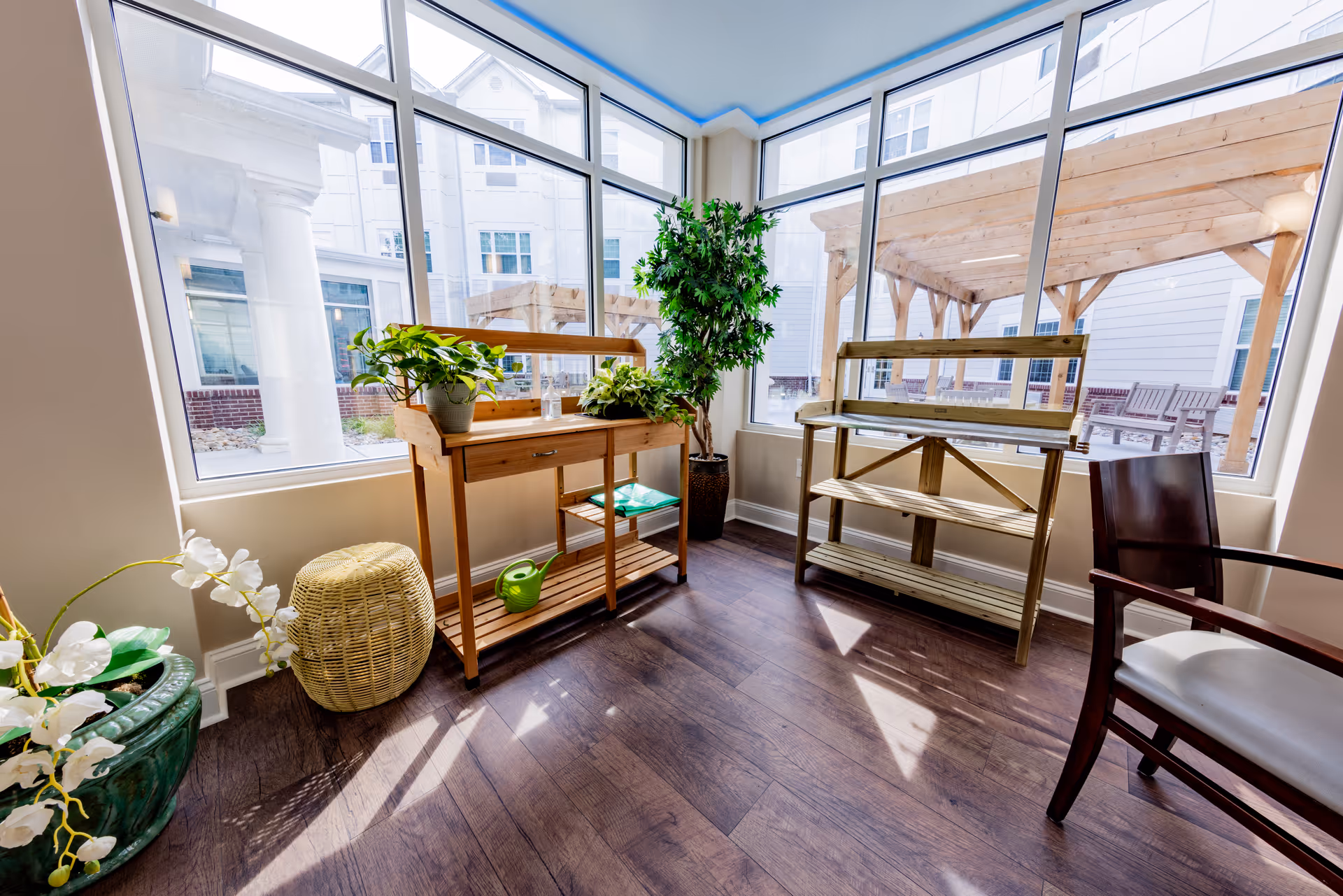 A bright indoor corner with large windows showing an outdoor patio. Inside, there are wooden plant stands with potted plants, a wicker basket, a green watering can, and a wooden chair with a white cushion. The floor is dark wood, and sunlight streams through the windows.