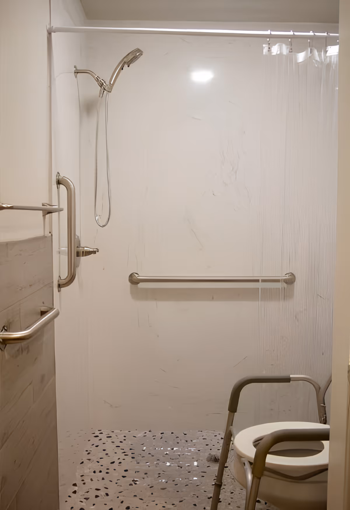 Accessible shower area with a handheld showerhead, grab bars on the wall, and a shower chair inside a walk-in shower with a clear shower curtain.