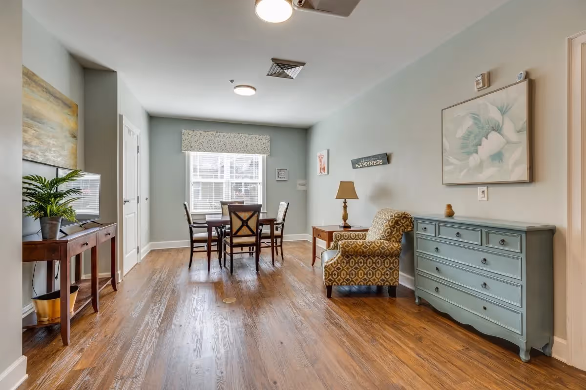 Bright communal room with a dining table and chairs, a patterned armchair, teal dresser, and a TV console with a plant.