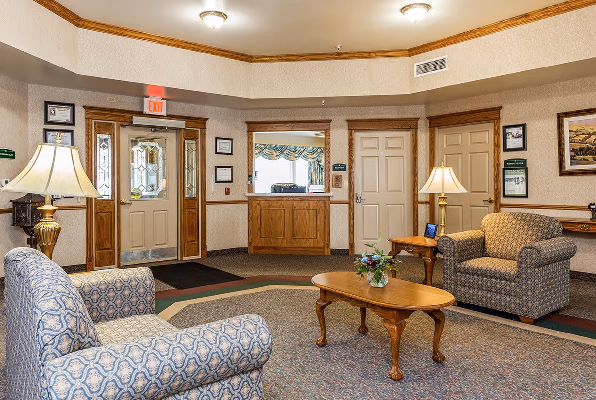 A cozy senior living facility lobby with two patterned armchairs, a wooden coffee table with a flower vase, two table lamps, and a reception window. The walls are beige with wooden trim, and there are several framed pictures and certificates. An exit door with glass panels is visible on the left side.