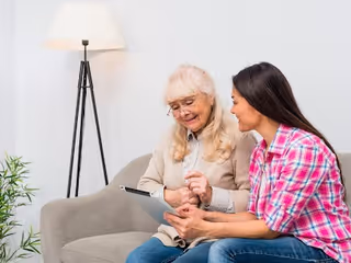 An elderly woman with long white hair and glasses sitting on a couch next to a younger woman in a pink plaid shirt. They are both looking at a tablet device and smiling. A tall floor lamp and a small green plant are visible in the background.
