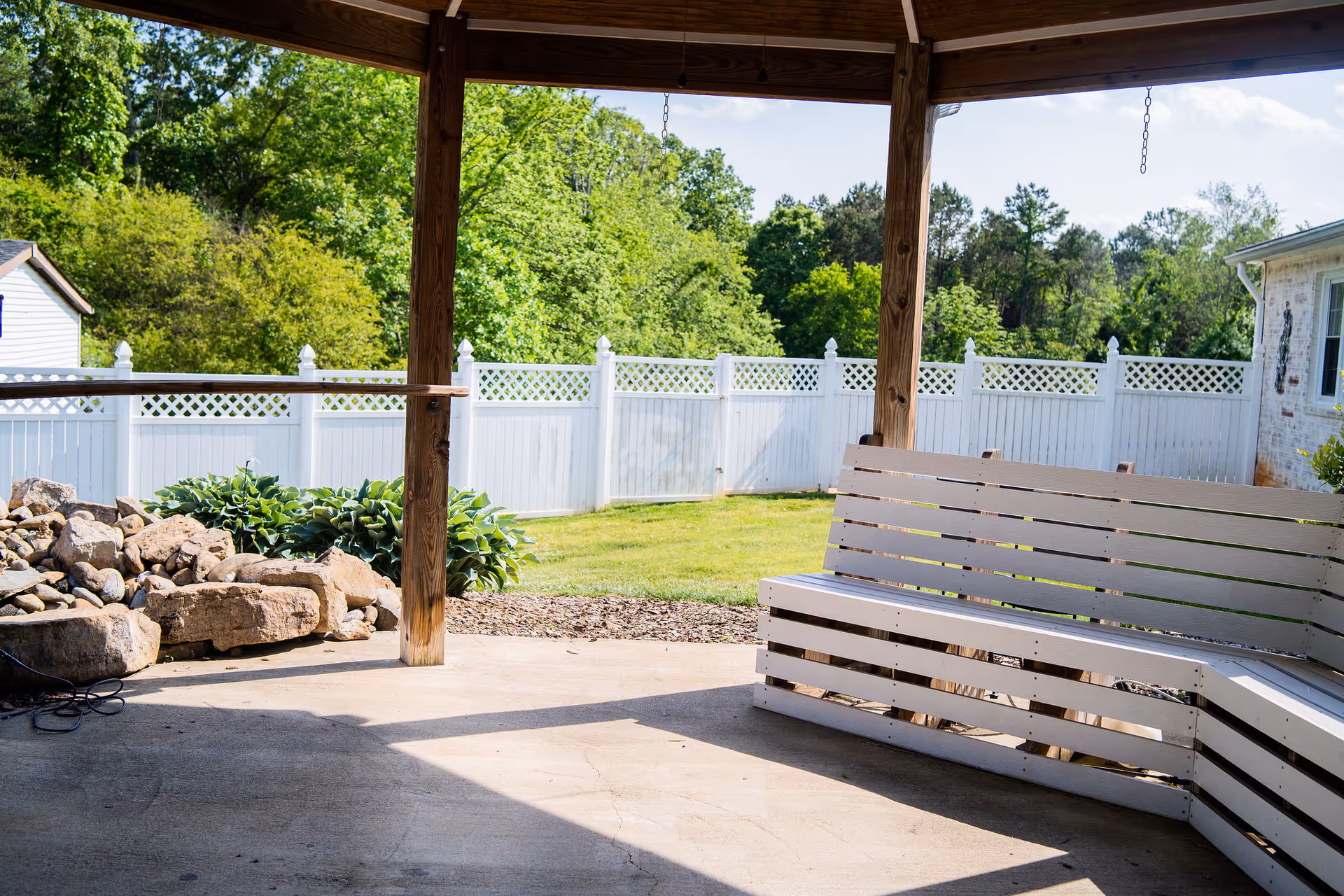 Covered patio with a wooden slatted bench swing, stone landscaping, and a white vinyl fence enclosing a grassy yard.