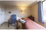 A small, simple bedroom in a senior living facility with a single bed covered in a pink quilted bedspread, a bedside table with a plant, a blue armchair, and a tray table holding a plate, cup, and utensils. A window with blue curtains allows natural light into the room.