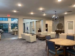 Spacious and well-lit common area in a senior living facility featuring multiple seating arrangements including white sofas, a round wooden table with chairs, and decorative elements such as a chandelier and wall art. Large windows and glass doors are visible in the background.
