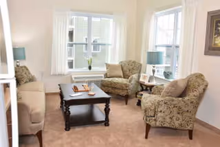 A cozy living room with beige carpet, two patterned armchairs, a beige sofa, a dark wooden coffee table with decorative items, two side tables with lamps, and large windows letting in natural light.