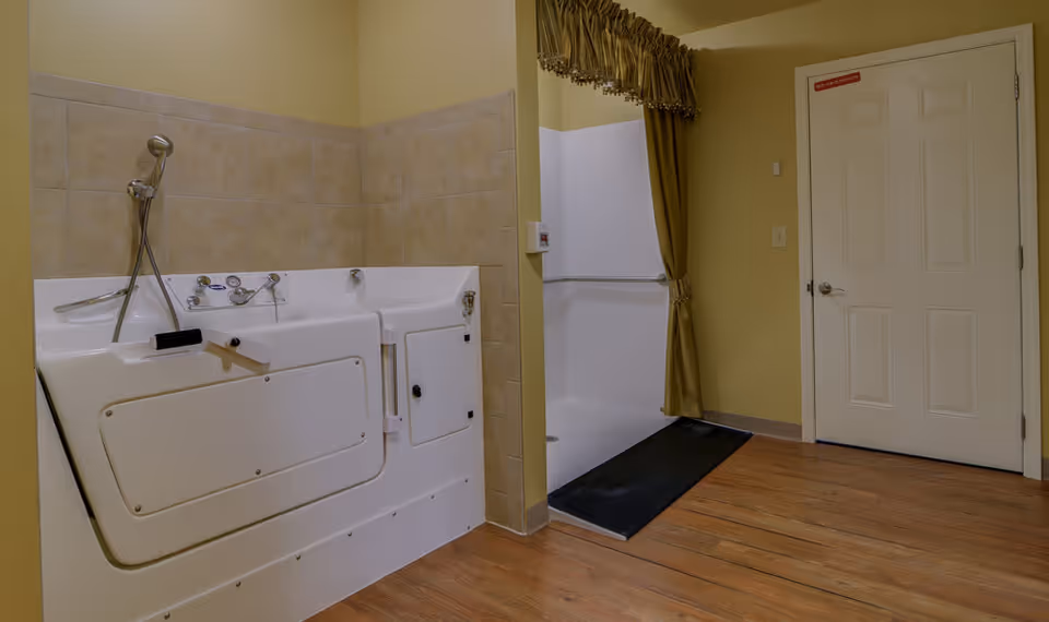 A senior-friendly bathroom featuring a walk-in bathtub with safety handles and a handheld showerhead. The room has beige tiled walls around the tub, light yellow painted walls, a wooden floor, a white door, and a curtained doorway leading to another area.