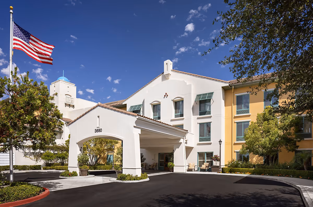 Exterior view of Belmont Village Thousand Oaks senior living facility featuring a white and yellow building with multiple windows, a covered entrance with the number 3680, an American flag on a flagpole, and landscaped greenery under a clear blue sky.