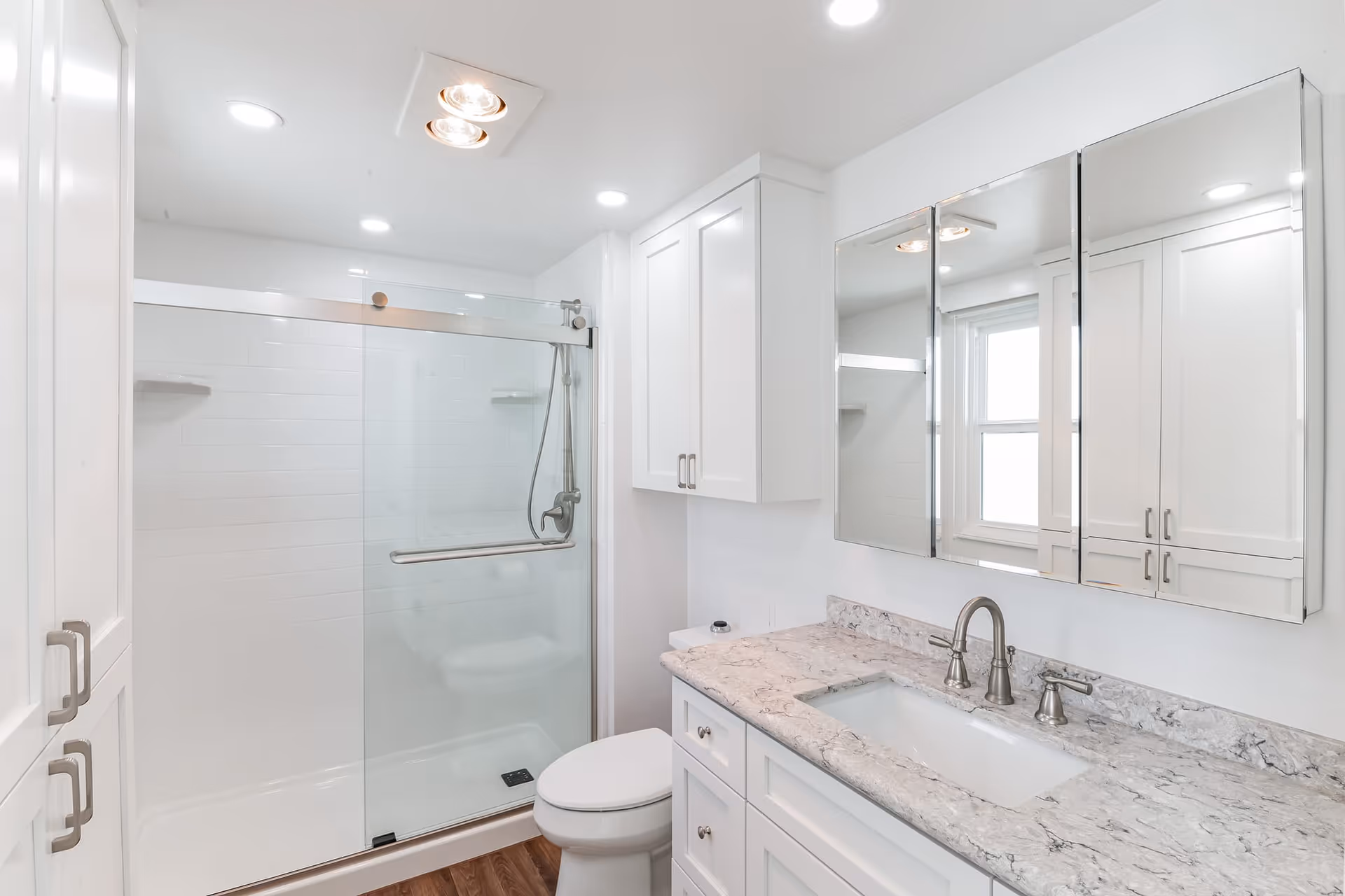 Bright and modern bathroom featuring a glass-enclosed shower with sliding doors, a white toilet, a marble countertop with an under-mount sink, and a large mirrored medicine cabinet above the sink. The room has white cabinetry and wood-look flooring.