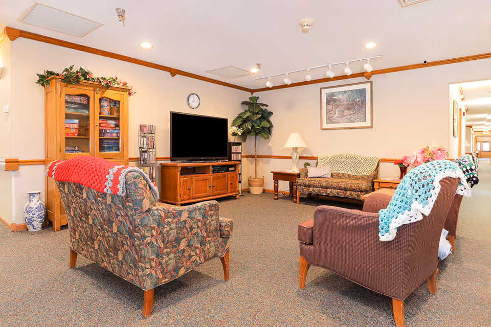 Common sitting area with upholstered armchairs and a sofa facing a television and wooden cabinet, decorated with lamps and plants.