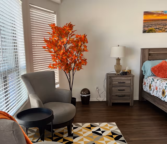 A cozy bedroom corner with a gray armchair next to a small black round table, a decorative artificial tree with orange leaves, a wooden nightstand with a lamp, a digital clock, and a tissue box. Part of a bed with floral bedding and a red pillow is visible. The room has wooden flooring, white walls, and windows with white blinds.