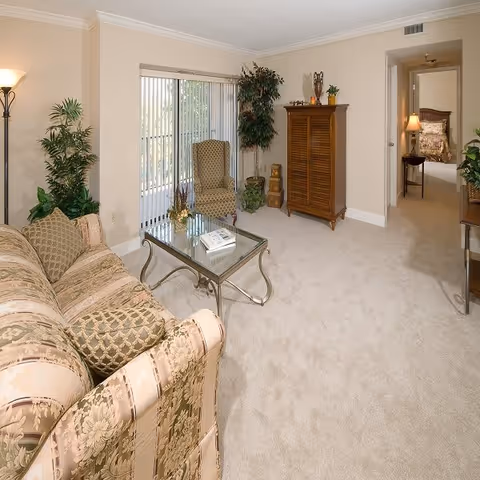 A cozy living room with a patterned sofa and matching armchair, a glass coffee table with books, potted plants, a wooden cabinet, and a sliding glass door with vertical blinds leading to an outdoor area. A hallway leads to a bedroom with a bed and nightstand visible.