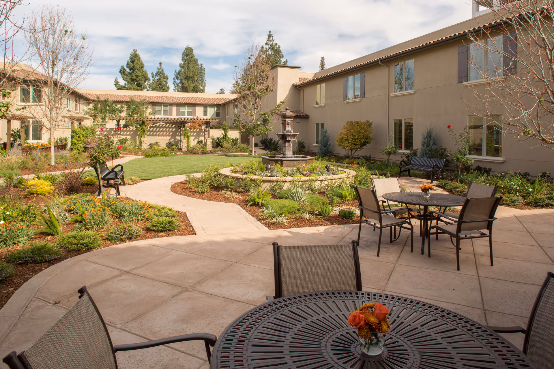 Outdoor courtyard area at Oakmont of Concord featuring a paved patio with round metal tables and chairs, each table decorated with a small vase of orange flowers. The courtyard includes landscaped garden beds with various plants and flowers, a central circular fountain, benches, and a two-story beige building surrounding the space under a partly cloudy sky.
