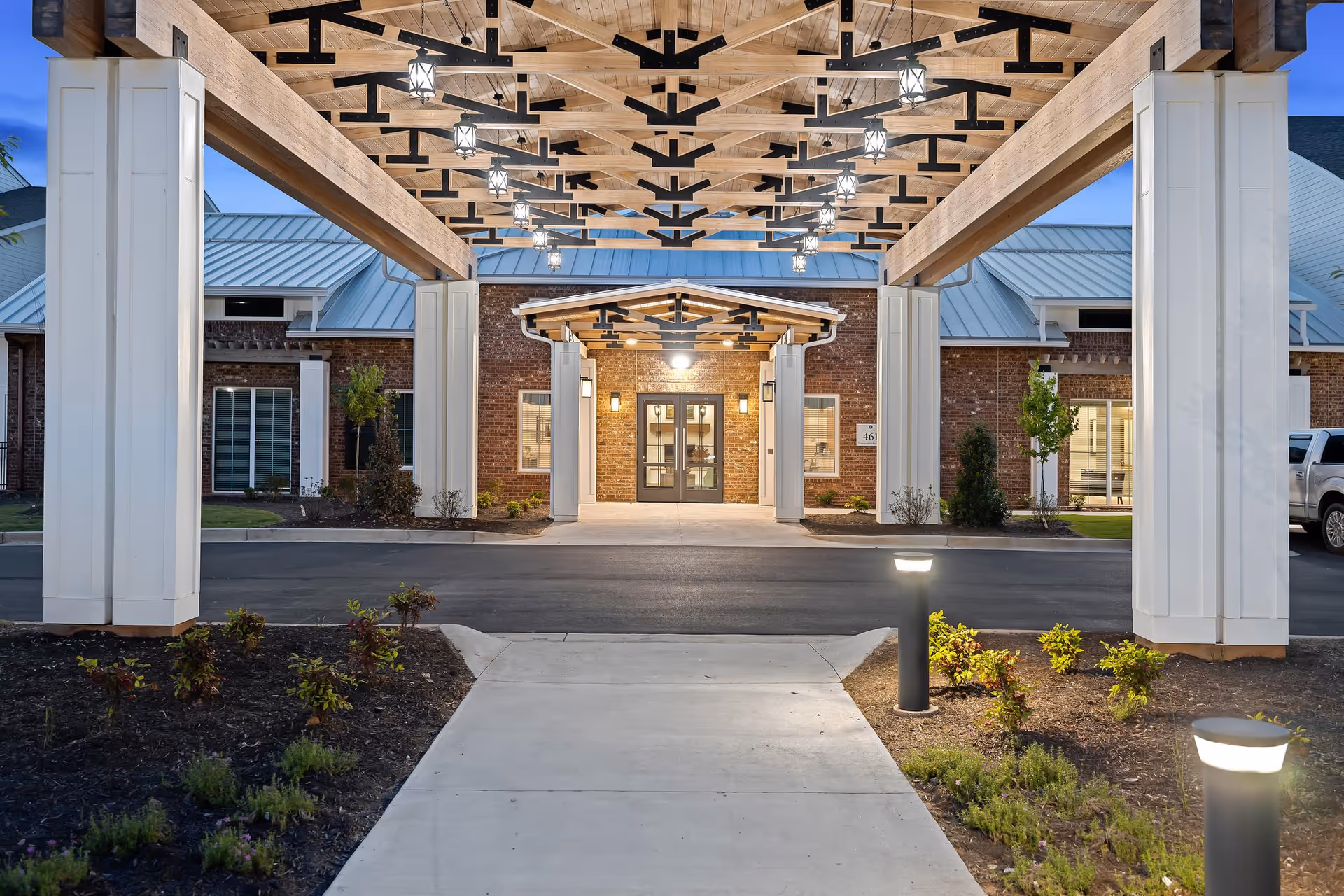 Entrance of The Claiborne at Brickyard Crossing senior living facility at dusk, showing a covered walkway with wooden beams and hanging lantern-style lights, leading to a brick building with glass double doors and windows on either side. There are landscaped plants and illuminated bollard lights along the concrete pathway.