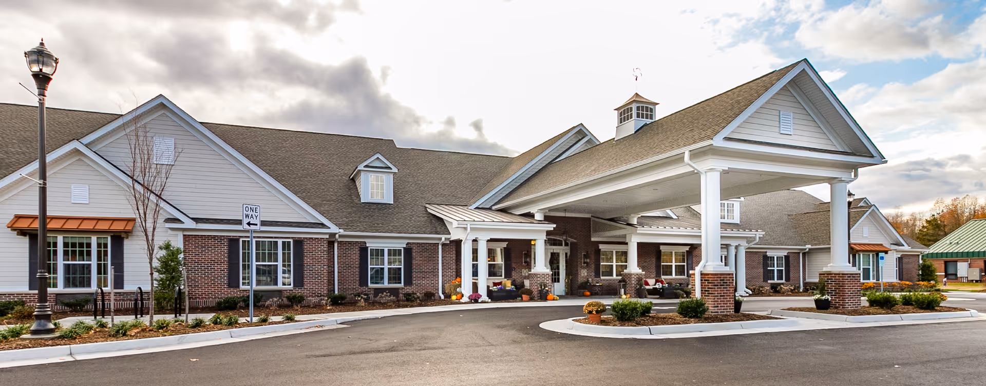 Exterior view of a senior living facility building with a covered entrance supported by white columns. The building has a combination of brick and light-colored siding with multiple windows and a small cupola on the roof. There are some plants and flowers near the entrance and a one-way street sign in front.